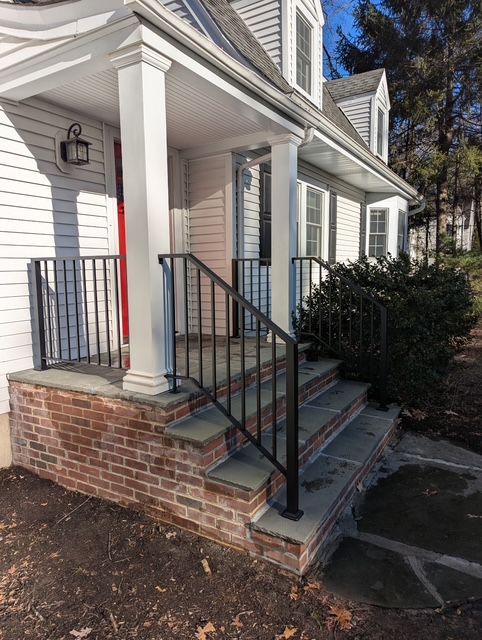 Front porch with brick steps, black handrails, white siding, and a small bush beside the entrance.