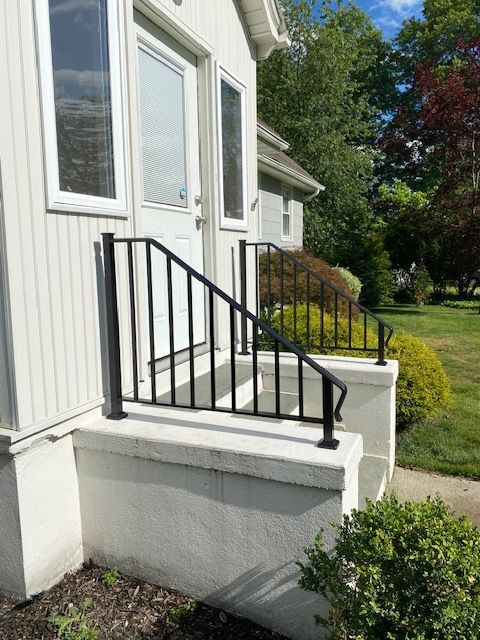 Front steps with black railing leading to a white house entrance, beside a landscaped yard.