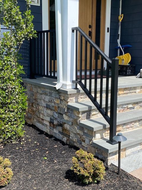 Front porch with stone foundation, black railing, gray steps, and a yellow broom near the door.