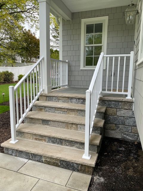 Front porch steps with white railings, gray siding, and stone foundation on a house.