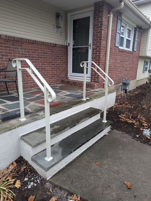 Front porch entrance with concrete steps, white railings, and a wheelchair ramp beside a brick house.