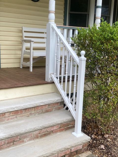 Front porch steps with white railing, wooden chair, and beige siding beside a shrub.