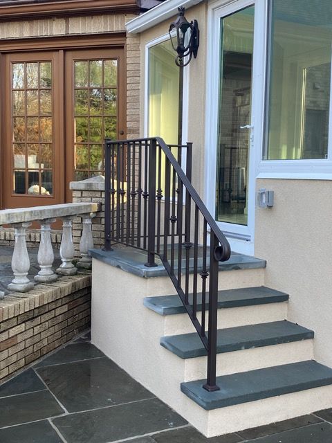 Front steps with black handrail leading to a house entrance beside a glass door and stone patio.