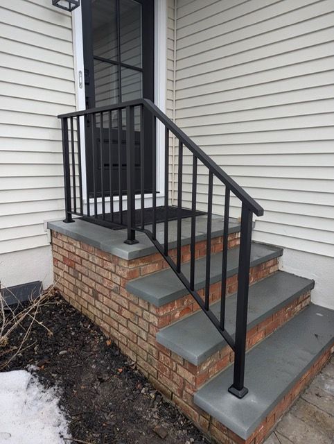 Front steps with black railing leading to a door beside a white house and brick foundation