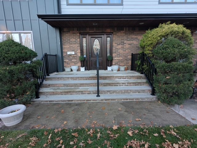 Front entrance with brick steps, black railings, double doors, and trimmed shrubs.