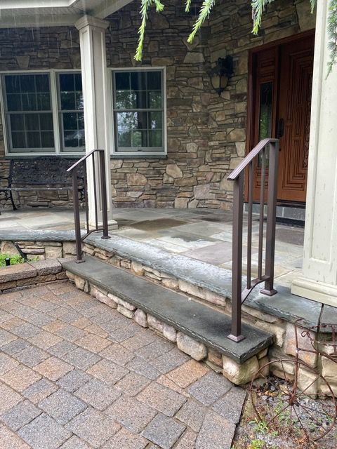Stone front porch with steps, black railings, and a wooden door beside windows on a brick walkway.
