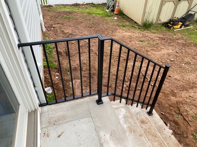 Black metal railing beside concrete steps on a small outdoor porch, next to a dirt yard and shed.