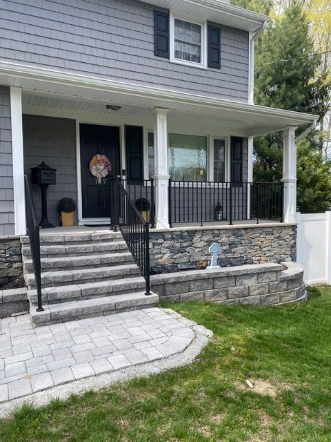 Front porch of a gray house with stone retaining wall, steps, black railing, and paver walkway.