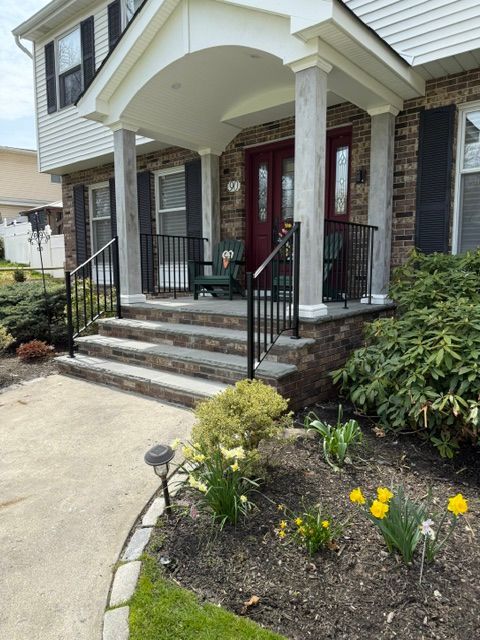 Front porch with steps, black railings, red door, and flower beds at a brick house