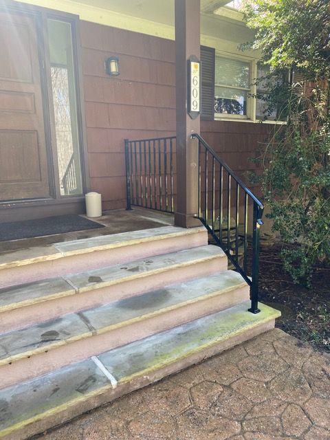 Front steps to a house with black railing, tan siding, and a small porch by a landscaped path