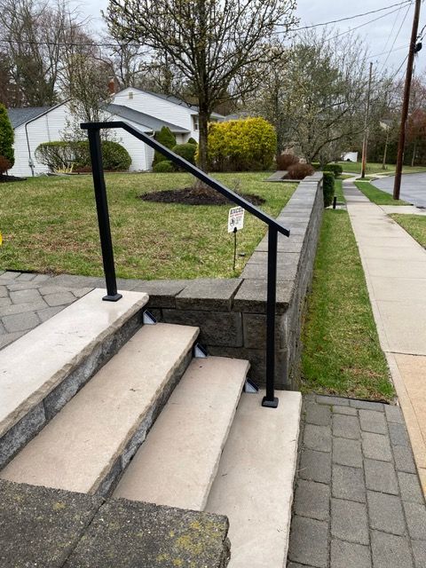 Front steps with black handrail leading up to a walkway beside a grassy yard on a residential street