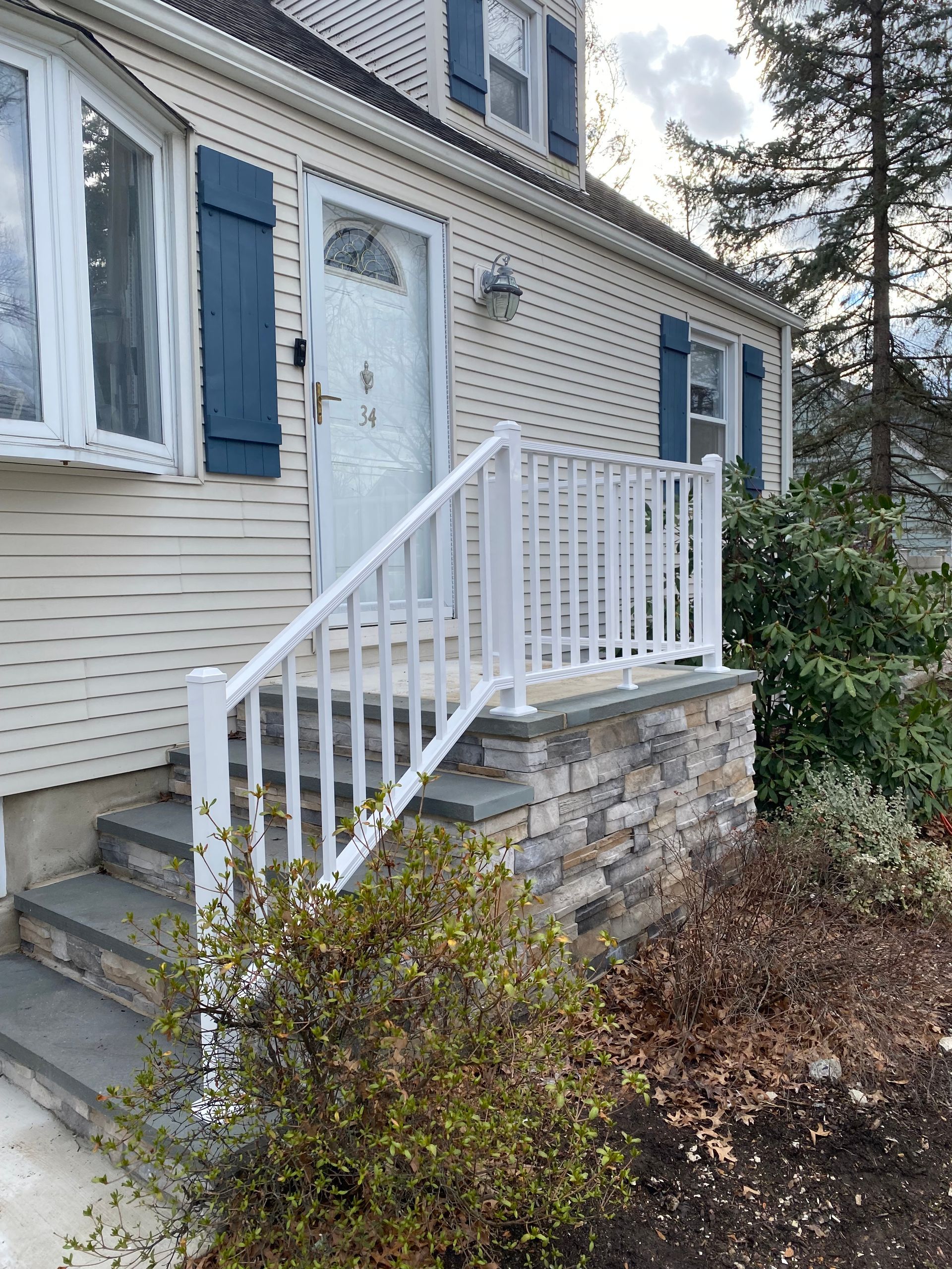Beige house with blue shutters and a white railing over stone facade steps leading to the front door.