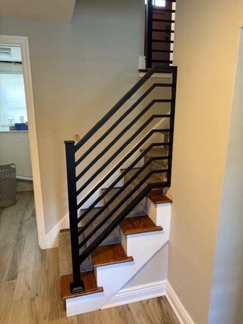 Staircase with horizontal black metal railing and wooden steps, beige walls, and wood-look flooring.