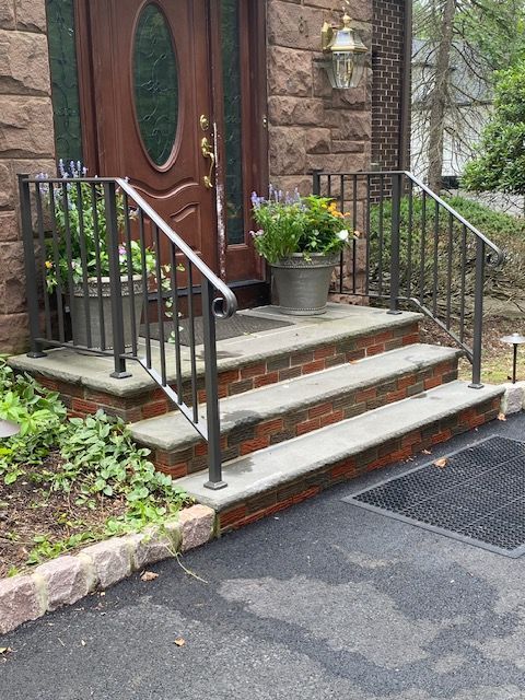 Exterior doorway with three brick steps, metal railings, and potted flowers.