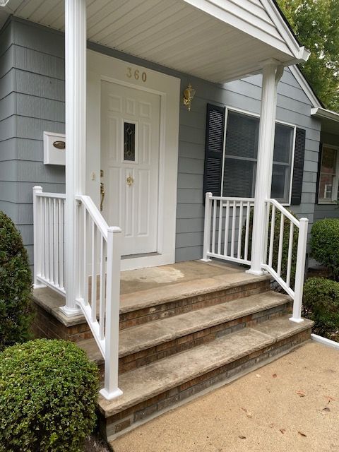 White-railed porch with three steps leads to a white door under a roof. Gray siding, shrubs, and a mailbox.