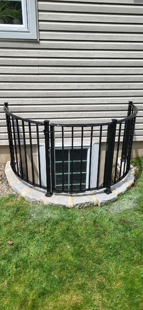 Black metal railing surrounding a basement window well on a beige house with green grass.
