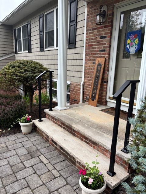 Black handrails on concrete steps leading to a house with brick and siding, potted plants on the porch.