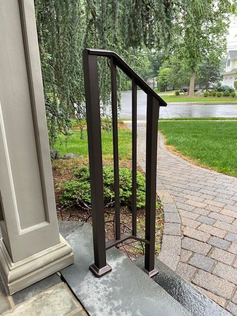 Brown metal handrail next to a gray stone porch, on a brick pathway, surrounded by green grass and trees.