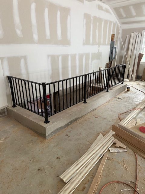 Black metal railing on a concrete step in a room under construction with drywall and lumber.
