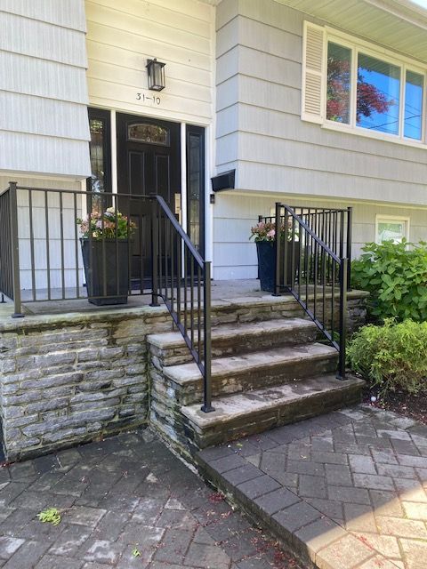 Gray house entrance with steps and dark railings. Steps lead to a black door with planters and a brick walkway.
