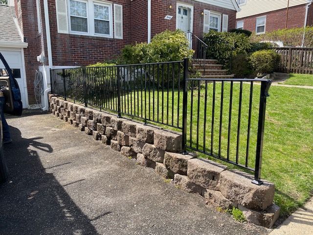 A black metal fence surrounds a stone wall in front of a brick house.