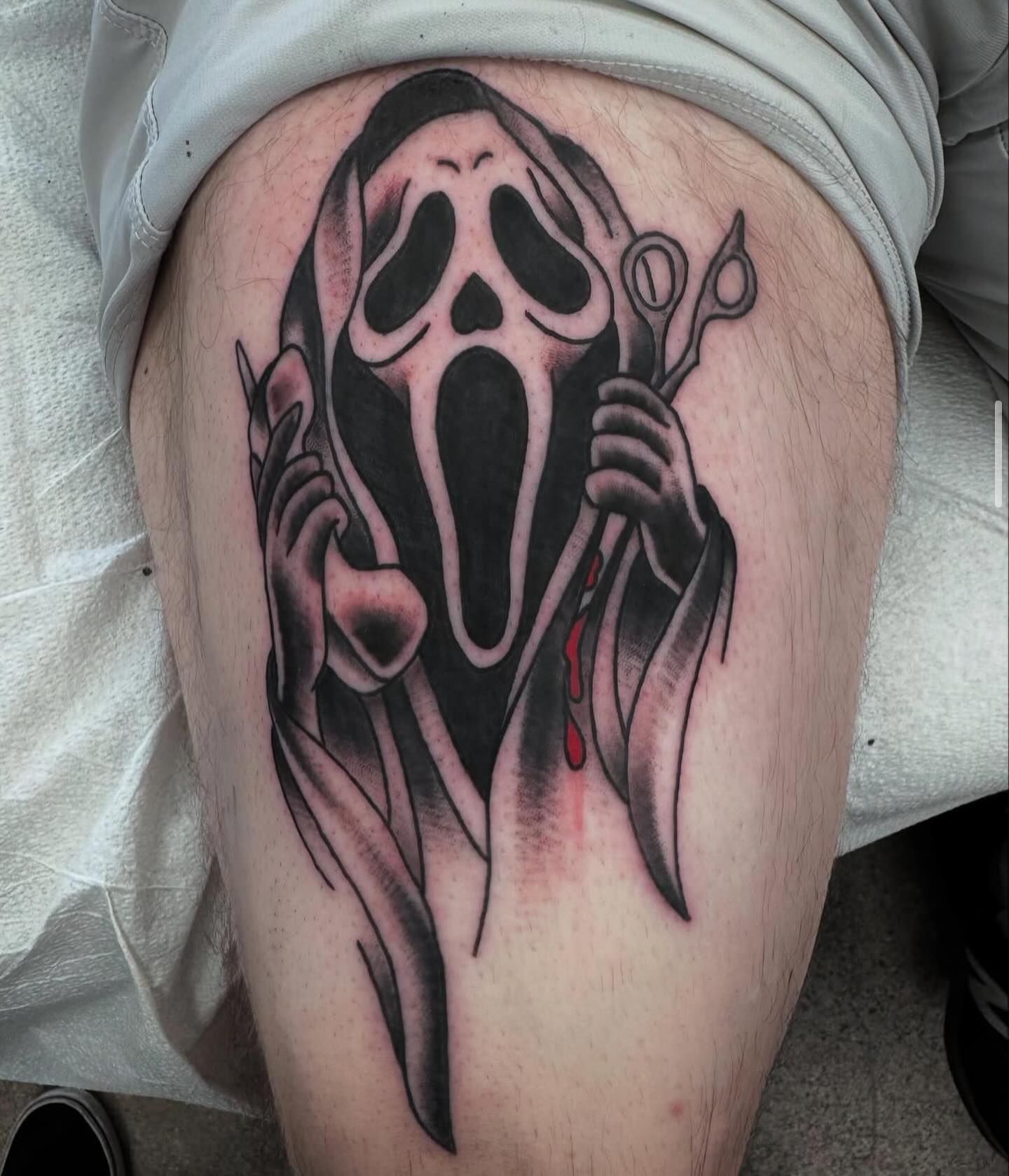 Tattoo of Ghostface from Scream holding scissors and a bloody flask on a person's thigh