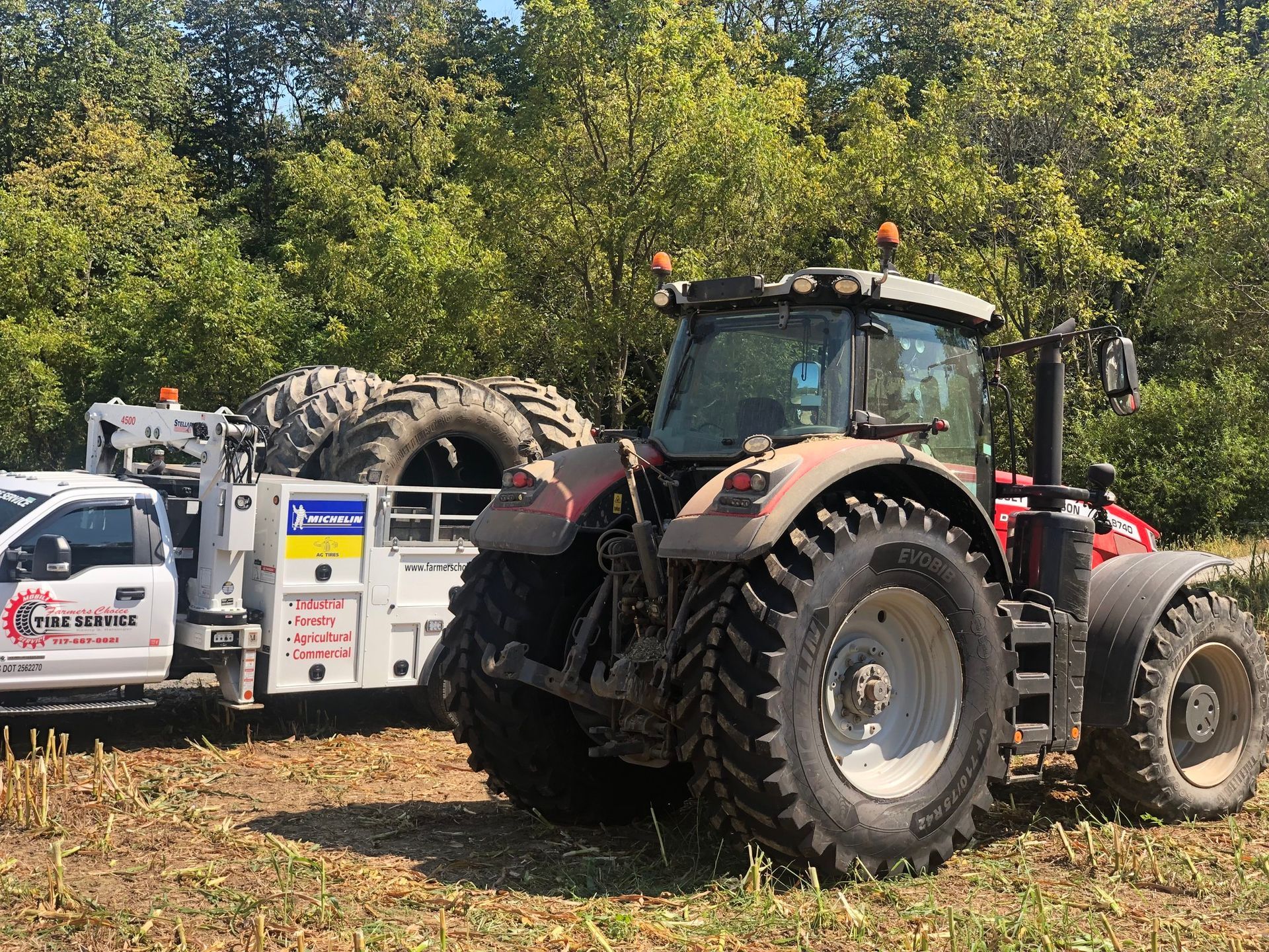 A red farm tractor parked next to a white service truck in a field, with large spare tires loaded on the truck bed.