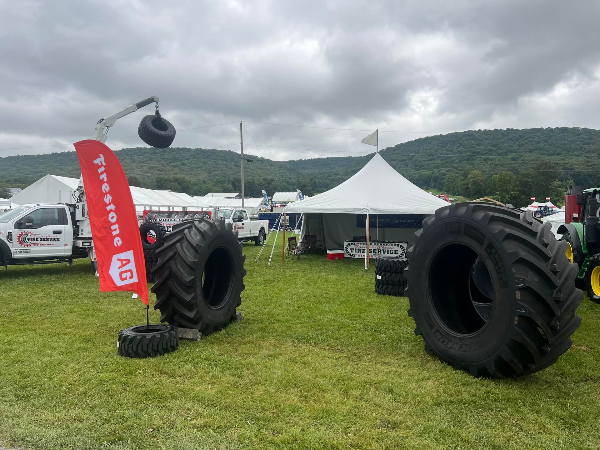 Large tractor tires on display at an outdoor event with a Firestone AG flag and tents against a mountain backdrop.