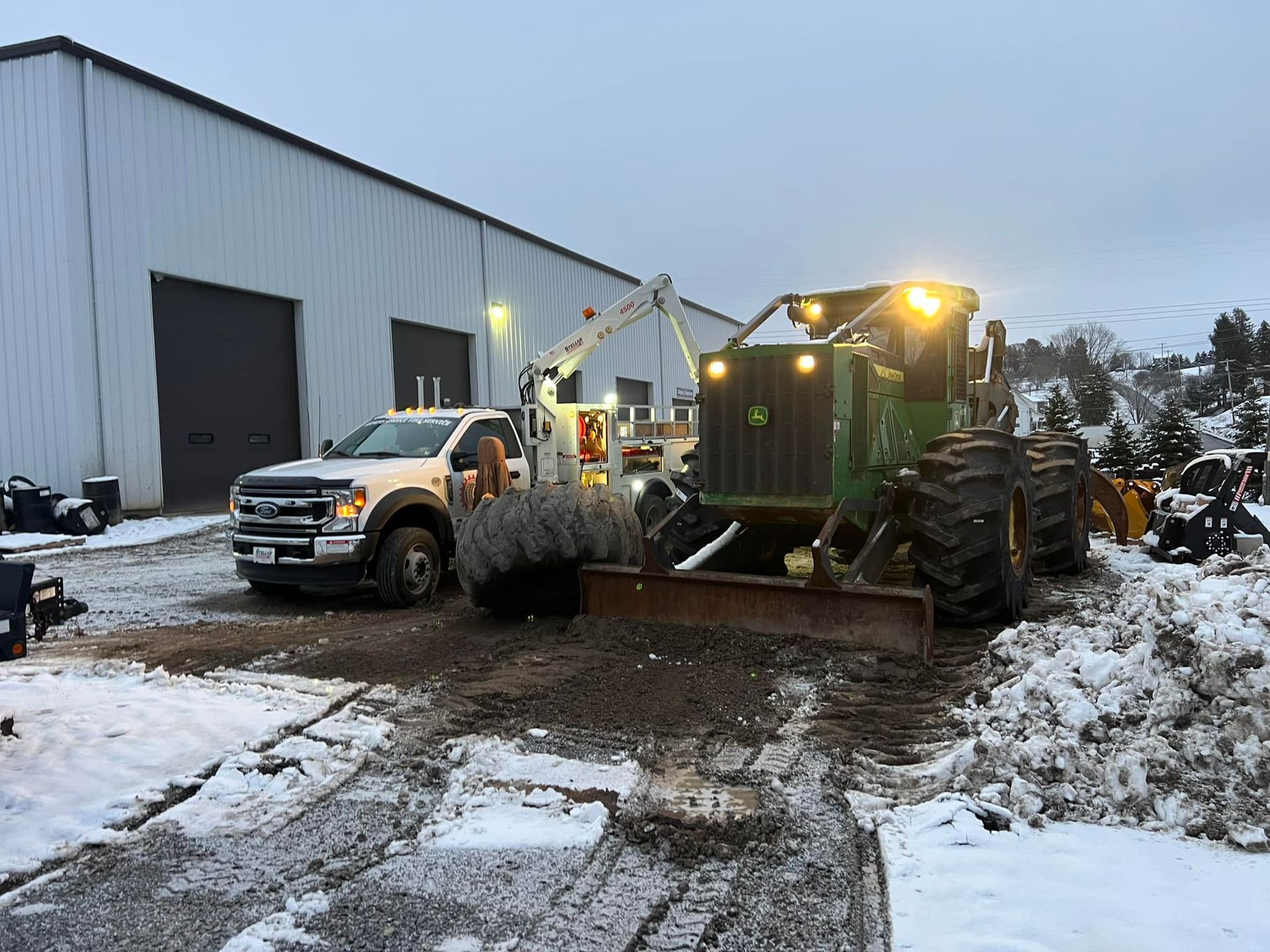 A white truck with snow plow and green tractor clearing snow in front of a building.