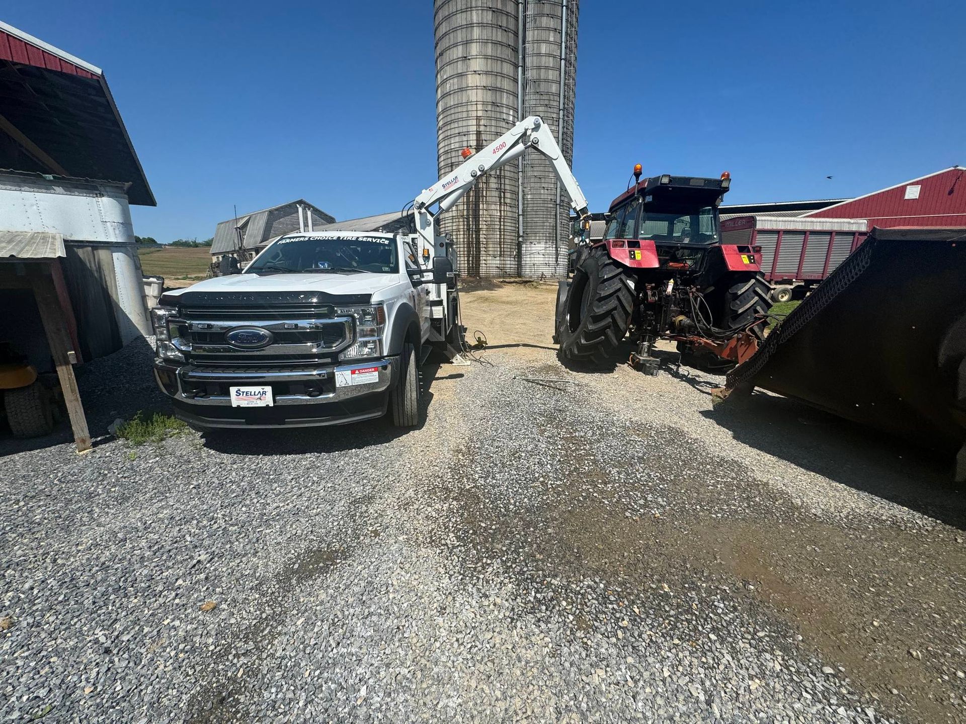 White truck with crane arm, red tractor, and silo in a gravel yard on a sunny day.