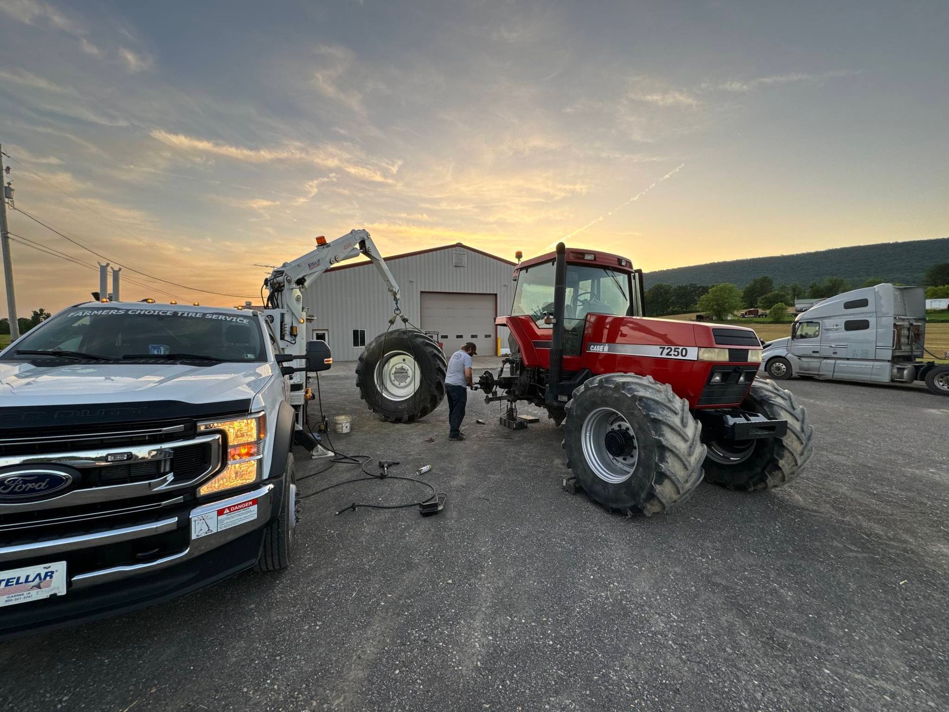 A red tractor with a flat tire being repaired, near a white truck and a garage at dusk.