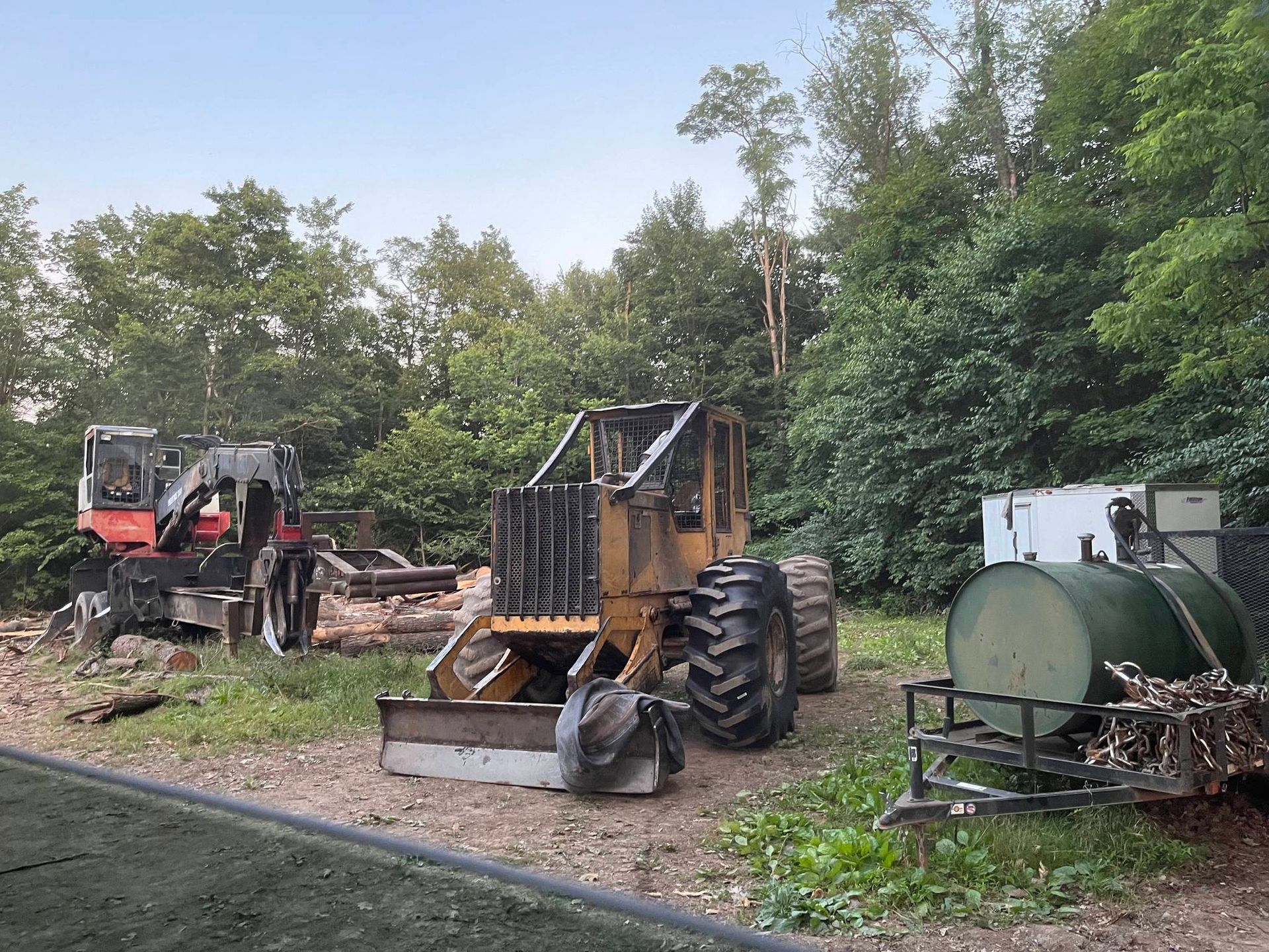 Two forestry machines on a clearing, surrounded by trees. A green tank and other equipment sit nearby.