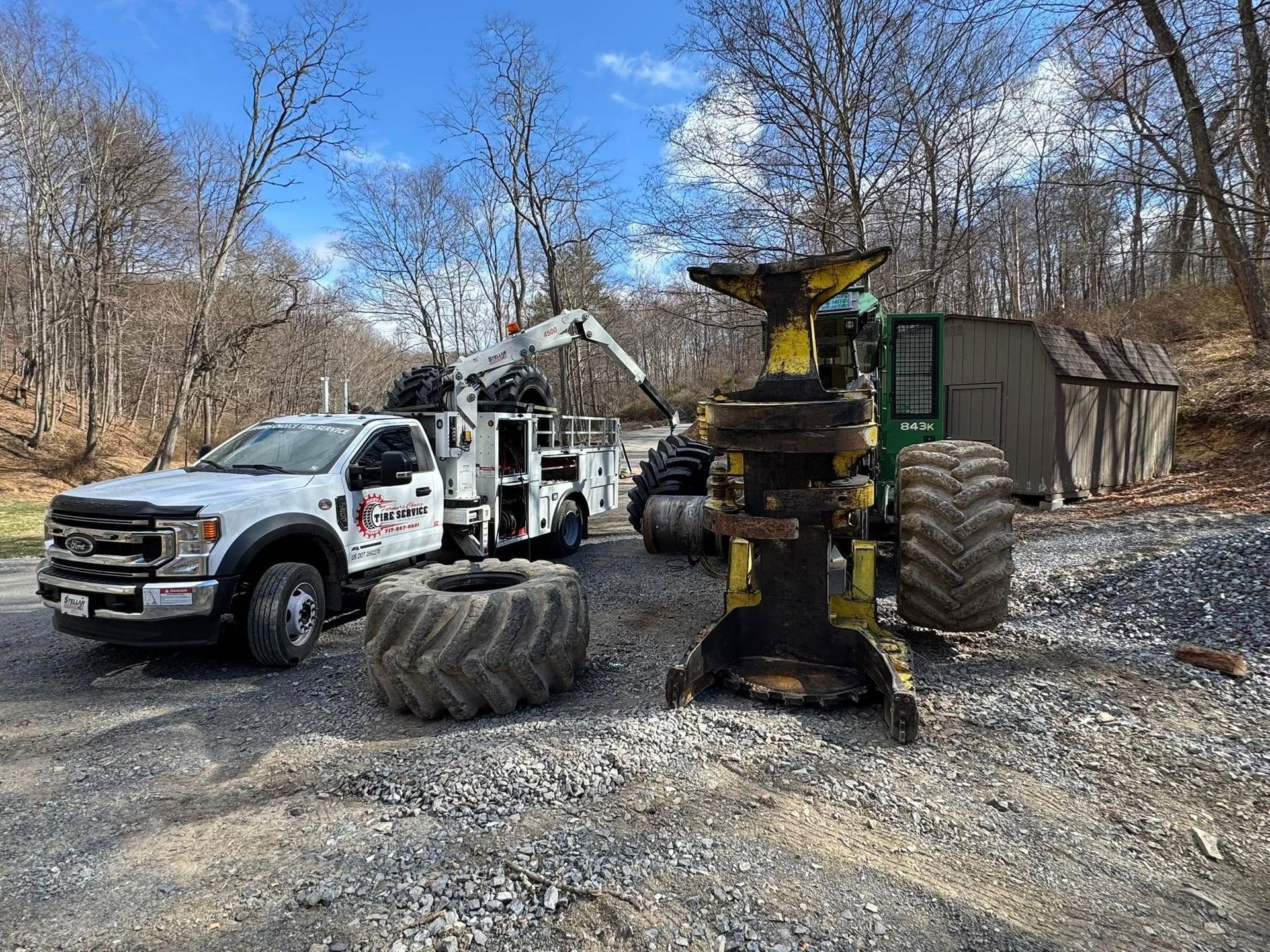A white truck with a crane, forestry equipment, and a large tire sit on a gravel surface.