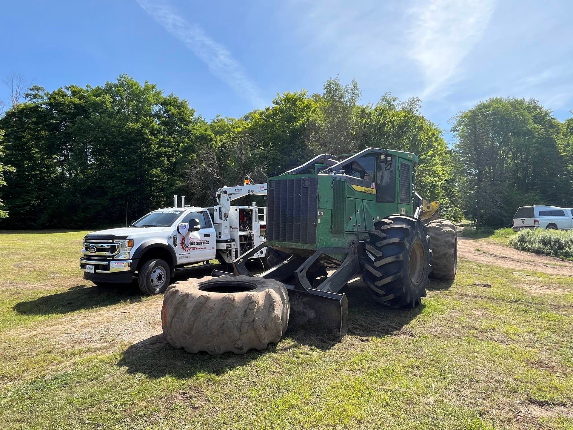 White truck with crane next to a green forestry machine on a grassy field; spare tire. Sunny day.