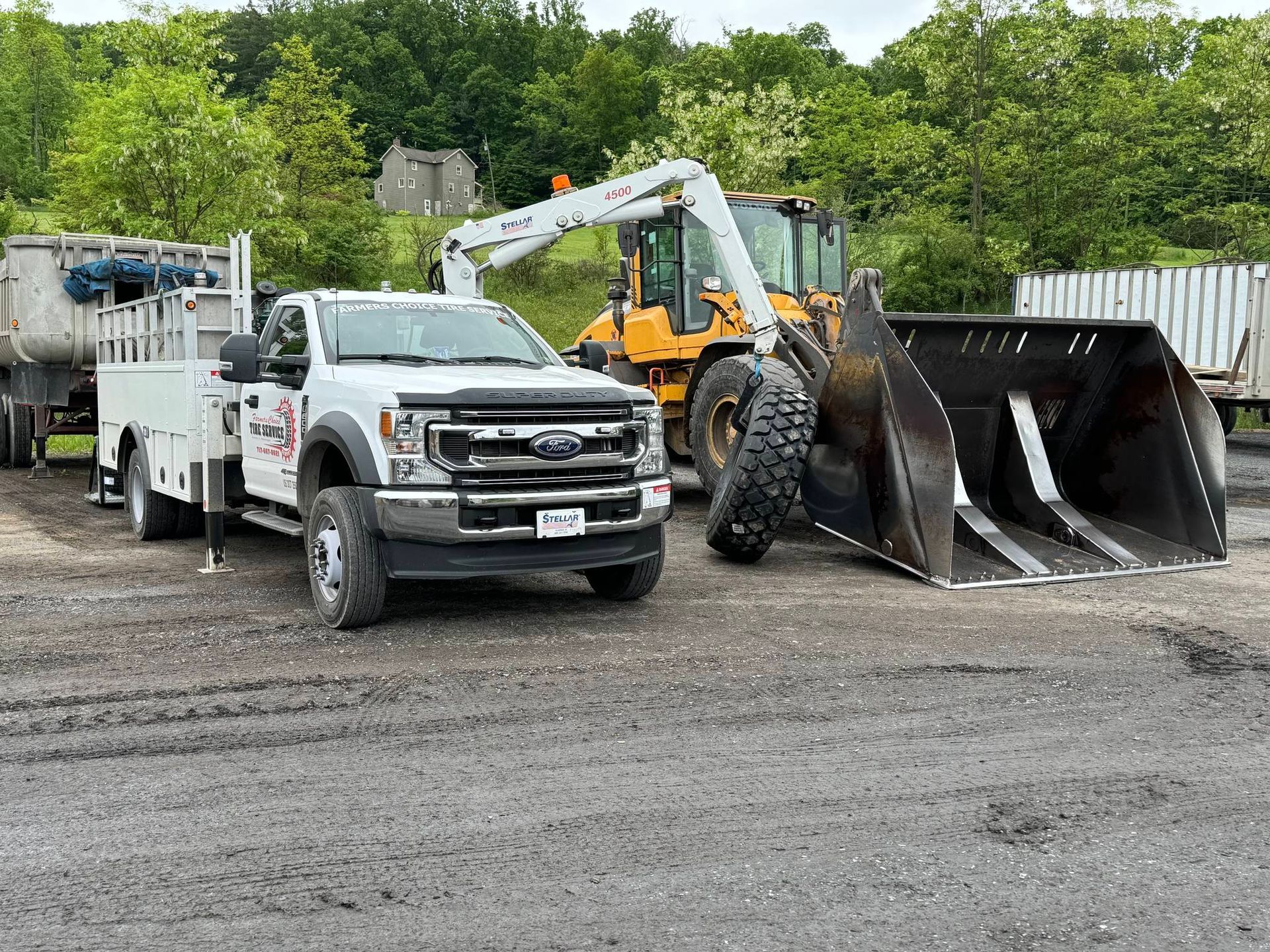 White truck parked next to a large excavator. Both are on a gravel lot near trees.
