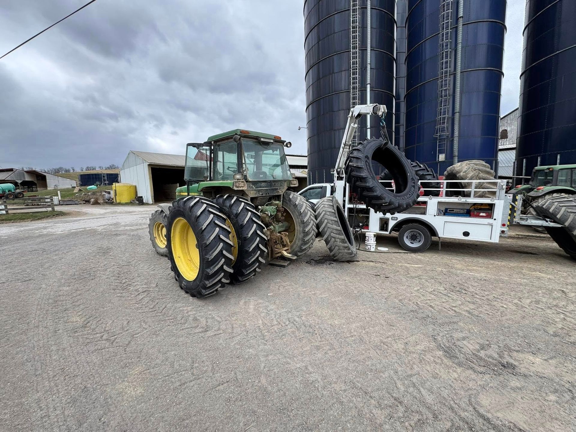 Green tractor with yellow wheels, parked on gravel next to silos and a utility trailer.