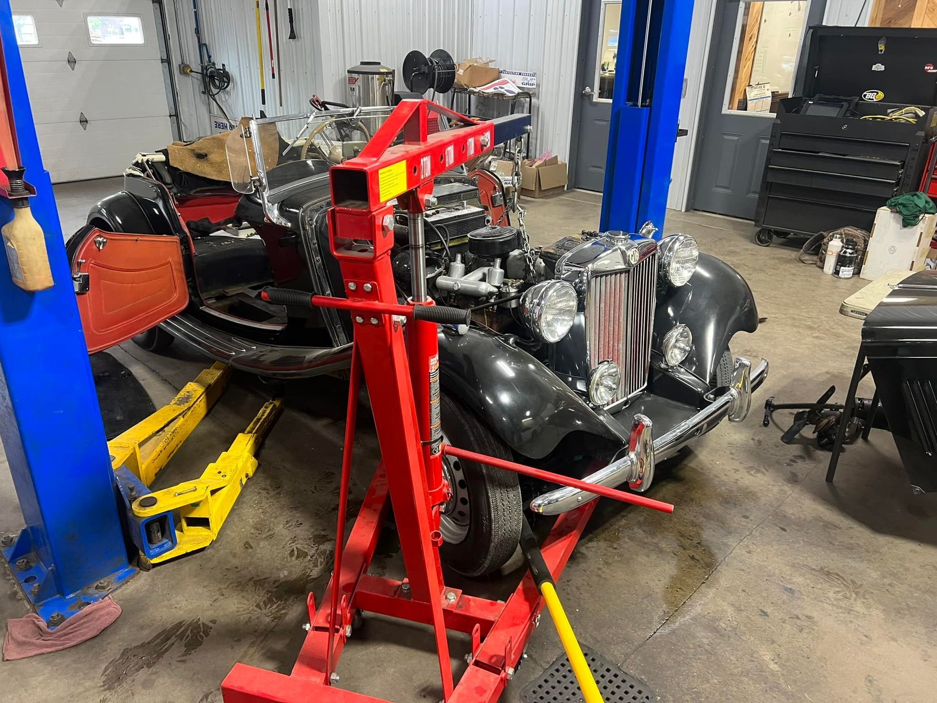 A red engine hoist sits in front of a vintage grey car inside a cluttered auto repair shop with blue lift columns.