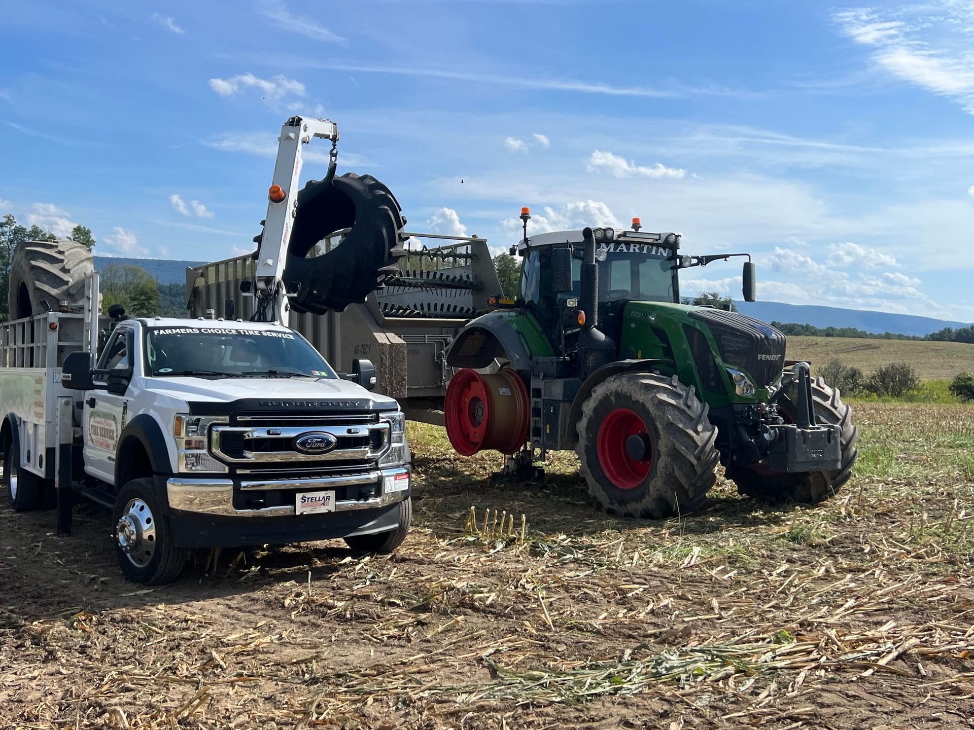 White truck with crane lifting a large tractor tire near a tractor in a field.