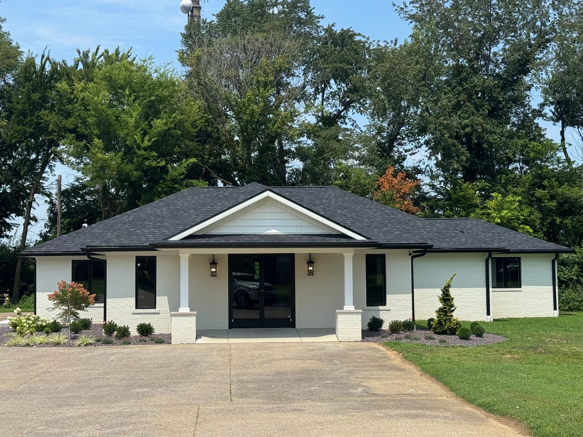 Single-story white house with dark roof, front porch, and a large driveway, surrounded by trees and lawn
