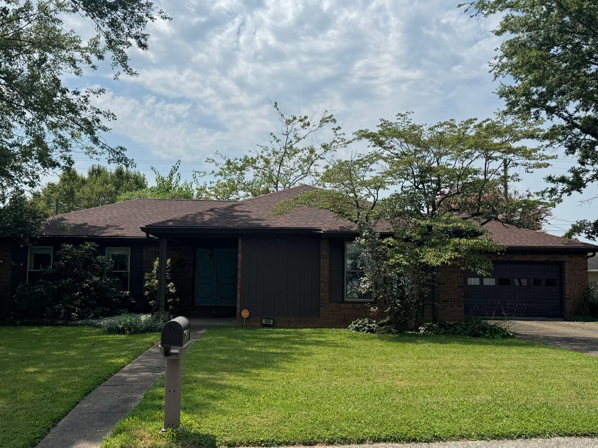 Single-story suburban house with a green lawn, mailbox, trees, and a garage under a cloudy sky