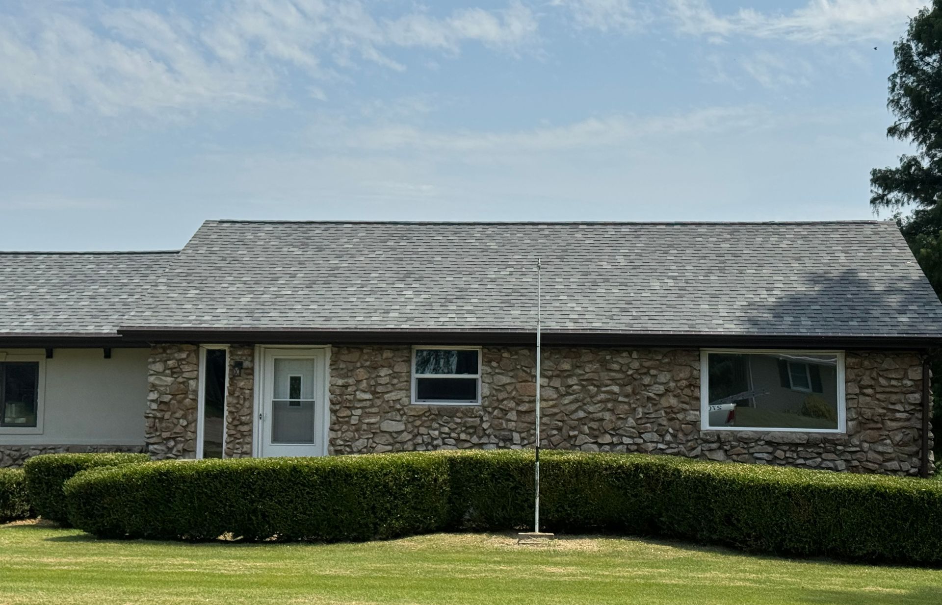 Single-story brick house with gray shingled roof, trimmed hedges, and green lawn under a blue sky