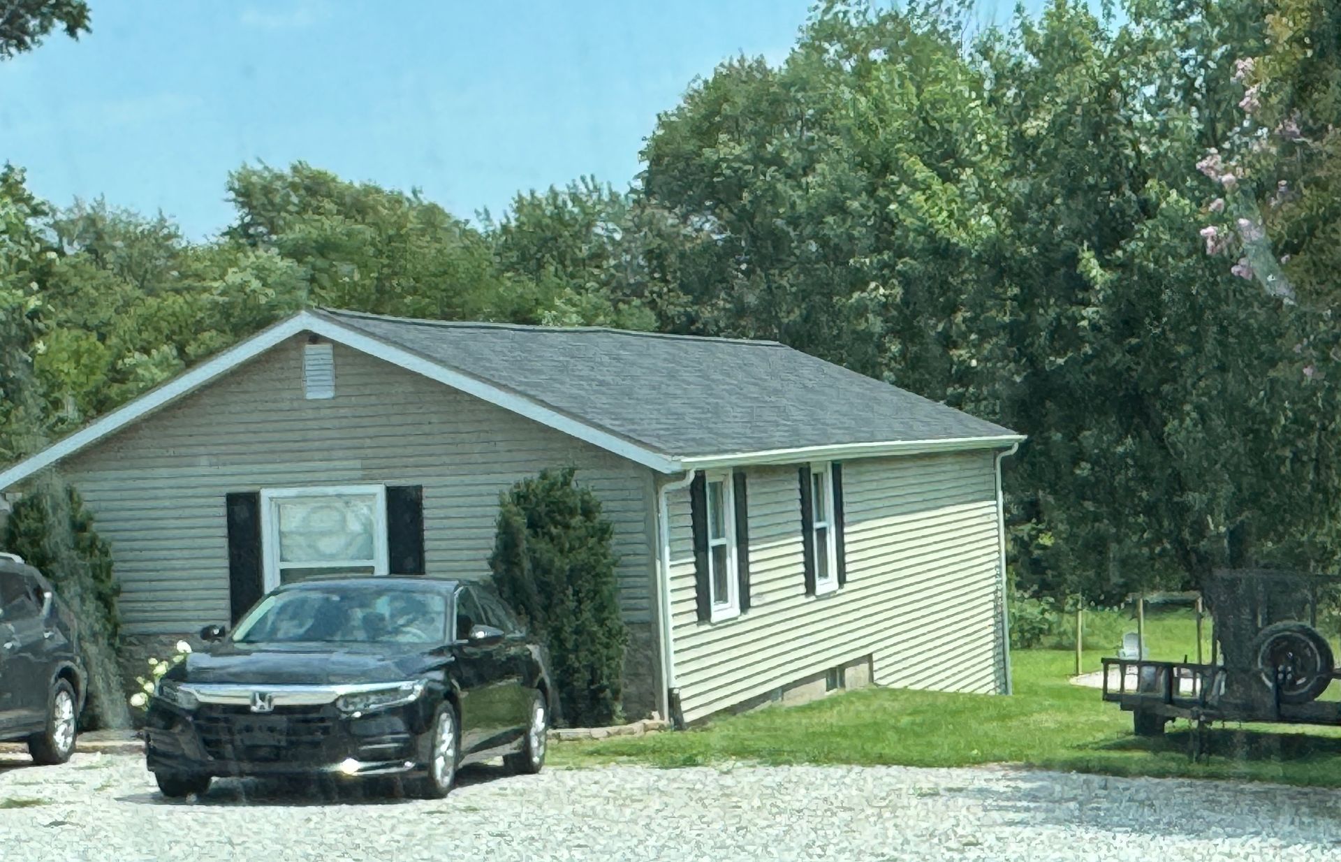 Single-story beige house with a black car parked in front, surrounded by trees and a gravel driveway