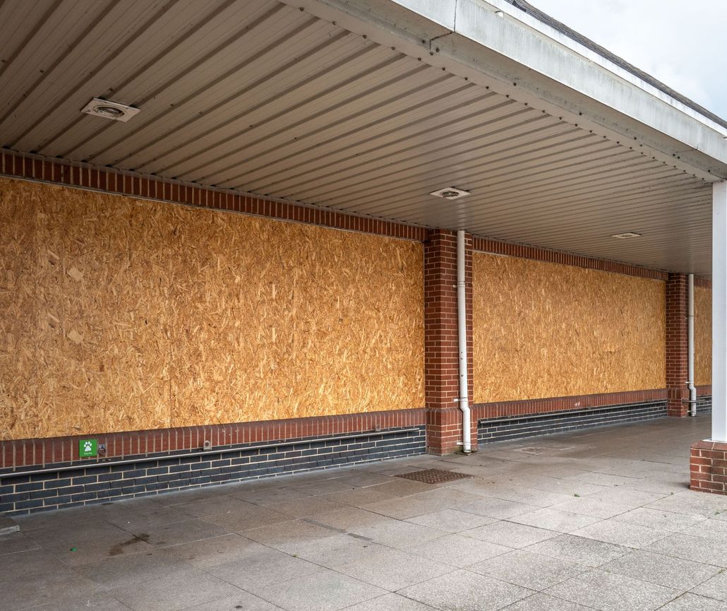 Boarded-up storefront with plywood-covered windows and brick columns under a covered walkway.