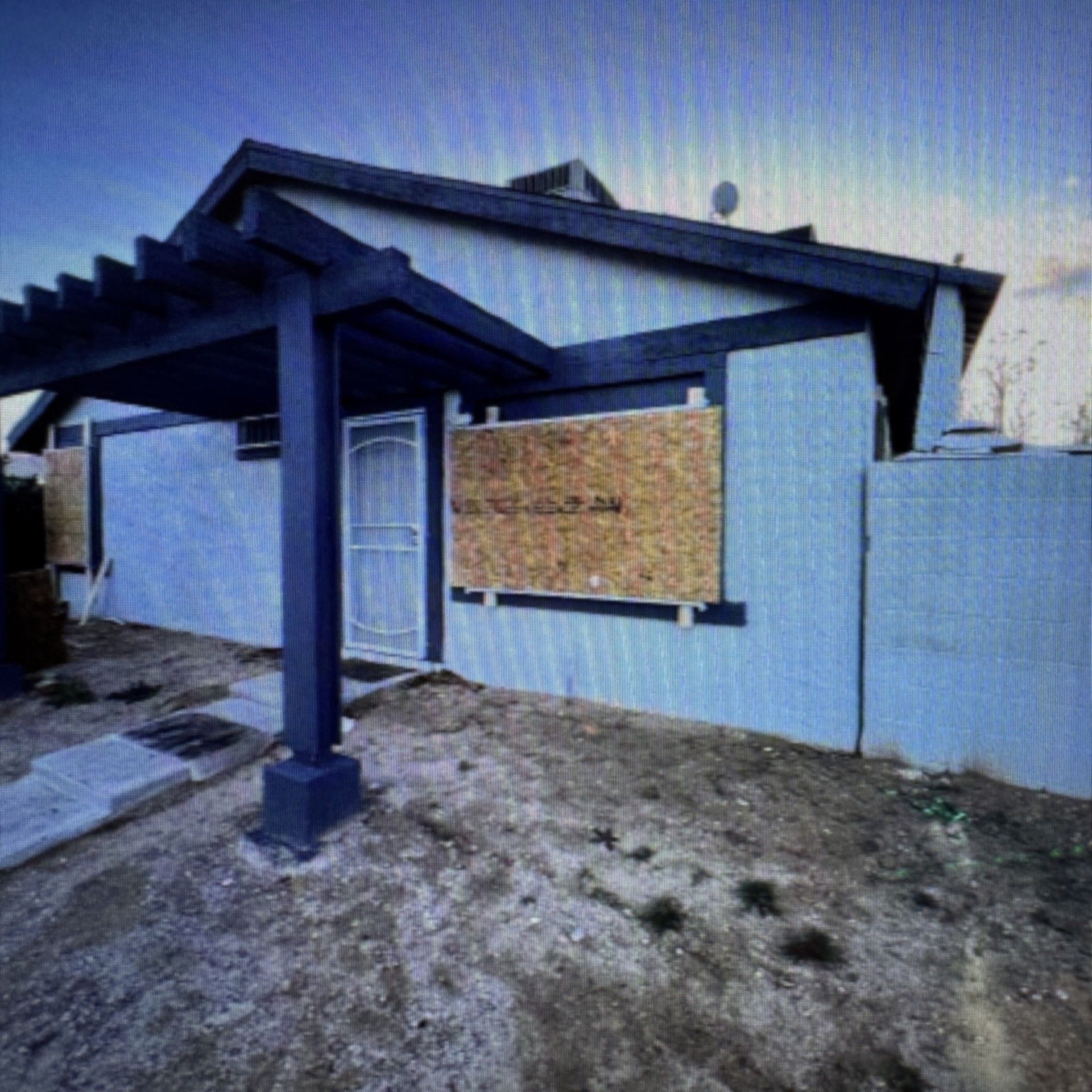 Boarded-up blue house entrance with a small covered porch and dry yard.