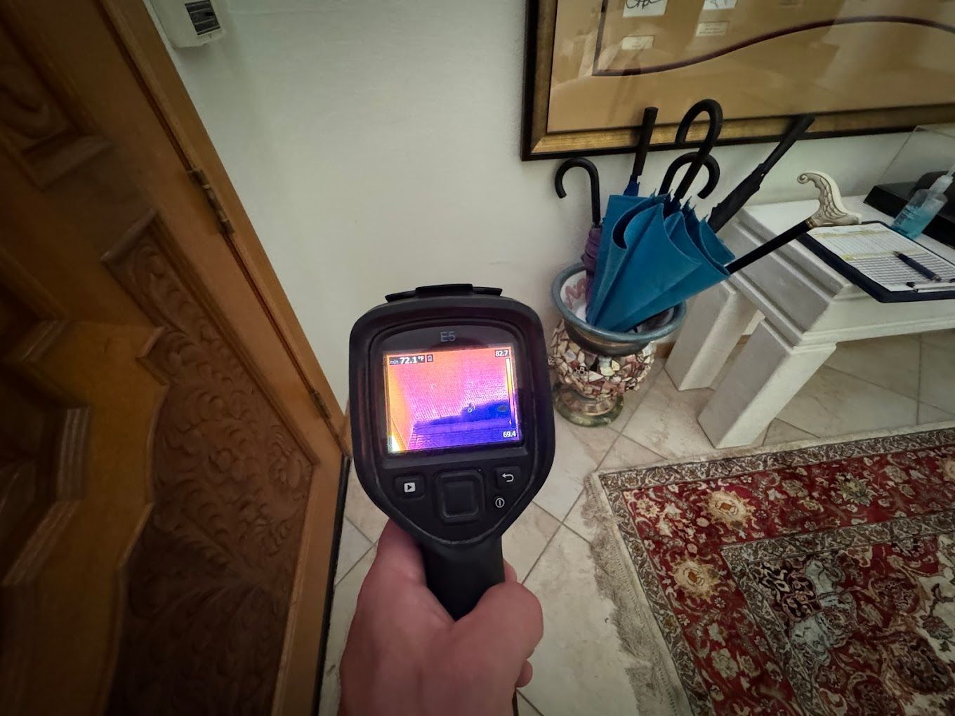 Handheld thermal camera showing a colorful heat map in a home hallway near a door and umbrella stand