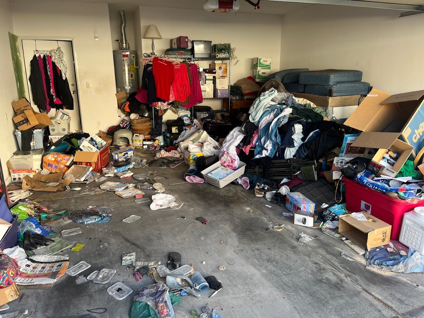 Cluttered garage filled with piles of clothes, boxes, papers, and scattered debris on the floor.