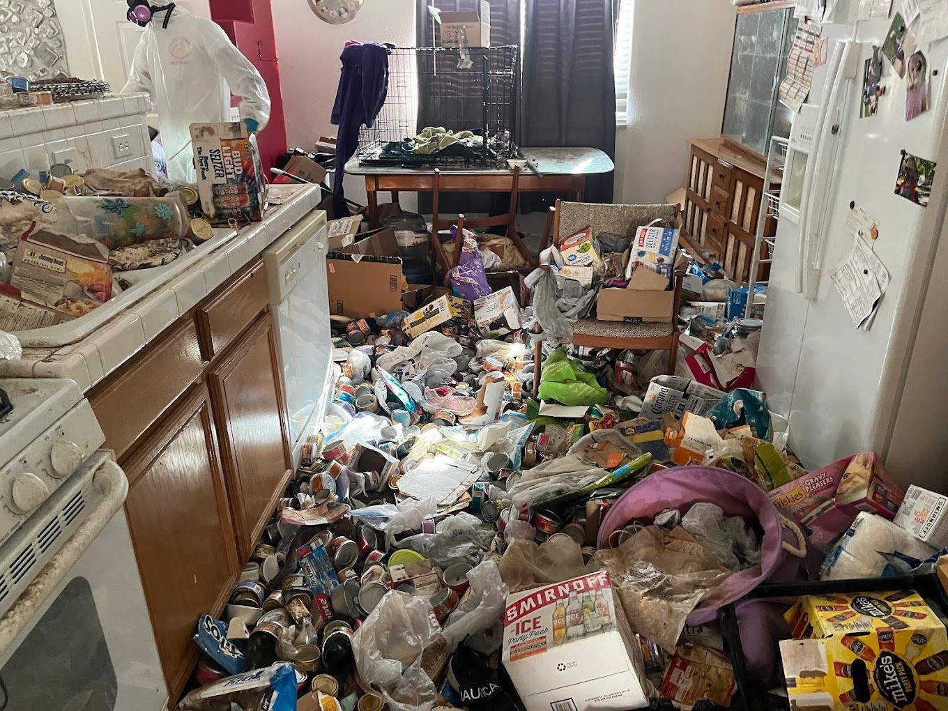Cluttered kitchen floor covered with trash, bottles, and debris, with cabinets and appliances along the sides