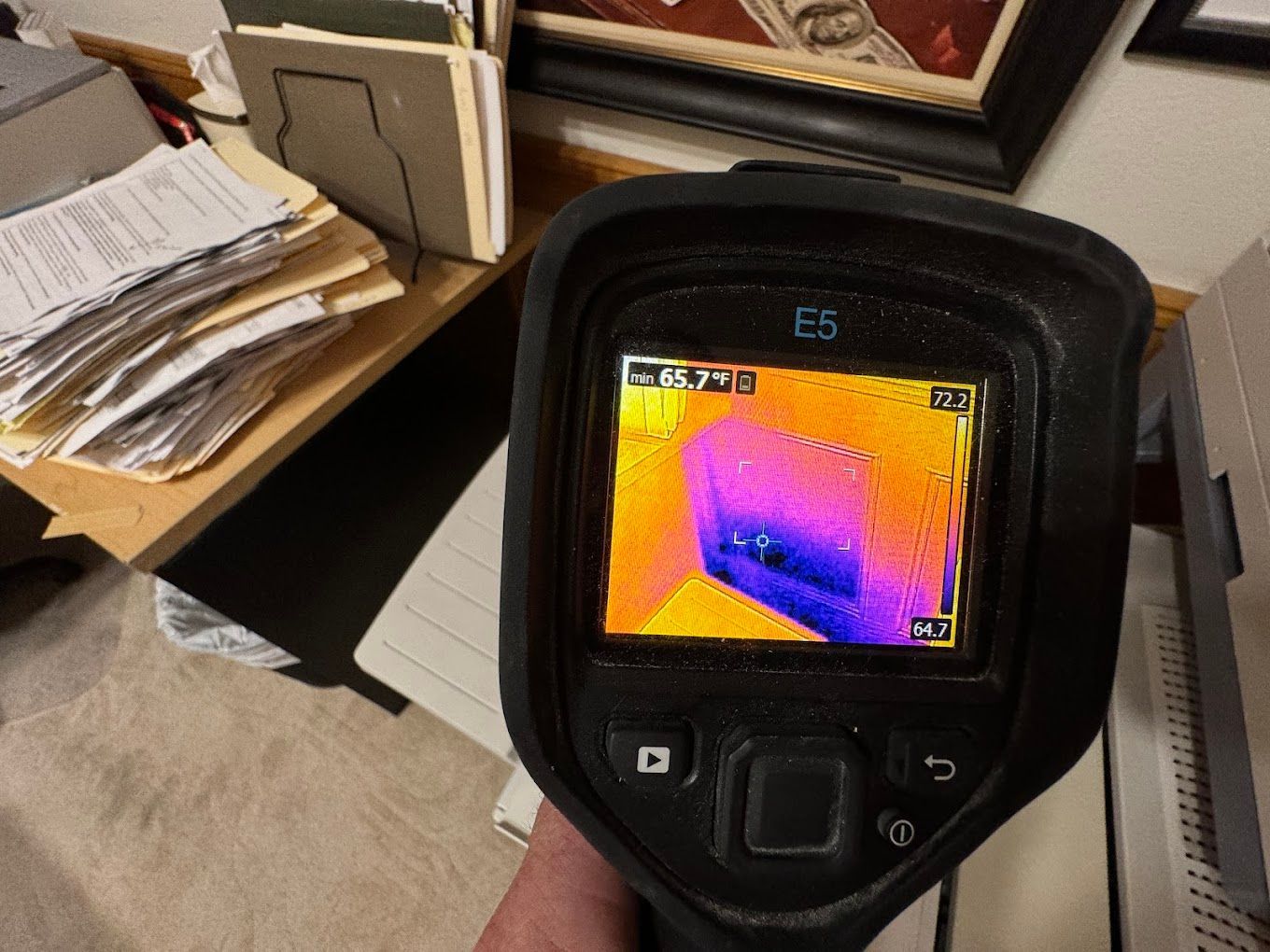 Handheld thermal camera displaying a colorful heat map on a table near papers and a laptop