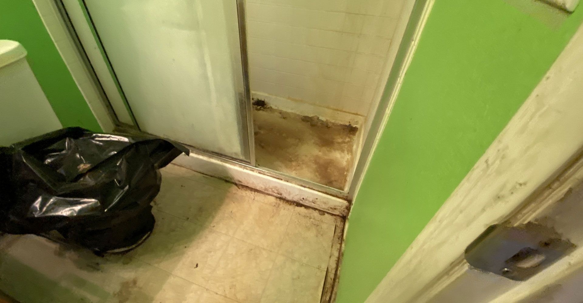 Narrow green hallway with a black trash bag on the floor and a dirty, damaged door at the end.