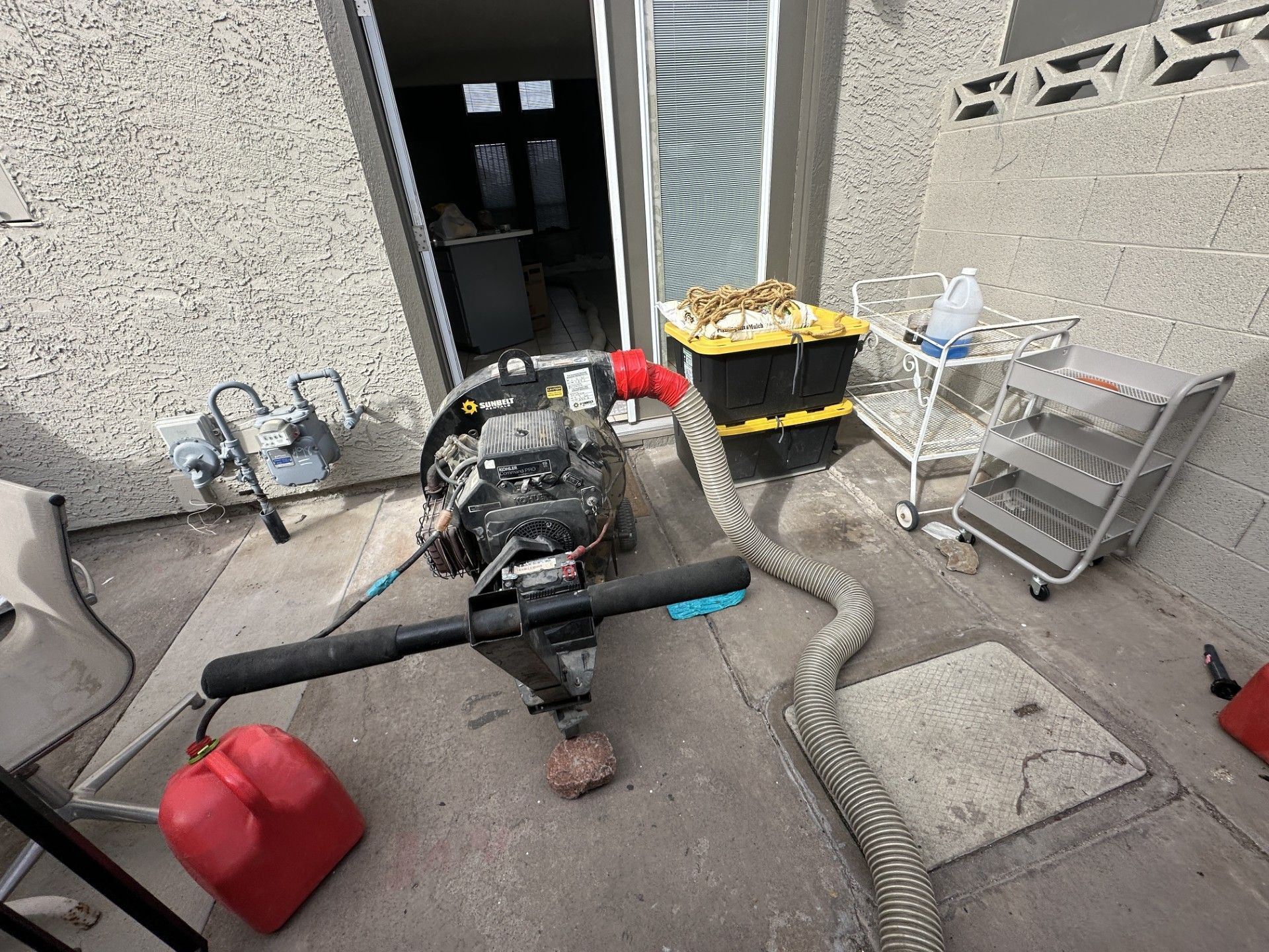 Outdoor utility area with a leaf blower, gas can, trash bins, and hoses near a building door.